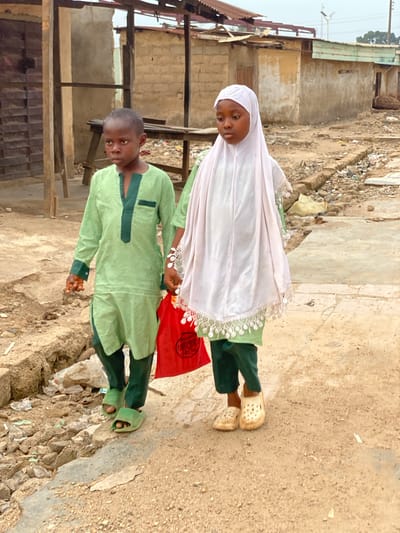 Two children, a boy and a girl, are standing in what appears to be an impoverished area in Kafanchan B, Nigeria. The girl wears a white hijab, a green dress, and light-colored clogs. The boy is wearing a green tunic-style shirt and pants with green sandals. They are standing on a rough dirt and concrete surface. Behind them is a dilapidated structure with a partially collapsed roof and exposed wooden and metal beams. Litter and debris are scattered around the scene. The overall environment suggests a low-income or slum area.