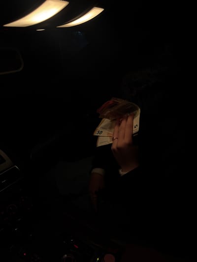 The image depicts a person holding a stack of euro banknotes in a dimly lit interior, likely the inside of a car, based on the visible dashboard elements. The hand holding the money wears a ring. The denominations of the visible banknotes include 5 and 50 euros. The location is not discernible as Valchedram, Bulgaria.