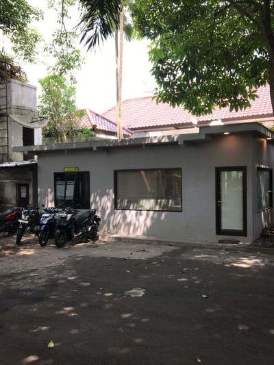 The image shows a small, grey building with a yellow sign that reads "Musholla" on the wall. There are two windows and a door on the front of the building. The windows have black frames and blinds. The door is also black and has a small window on the top. The building is located on a paved driveway with a few motorbikes parked in front. The driveway is shaded by a large tree with green leaves. The location is in Malang, Indonesia, at the coordinates -7.98 latitude and 112.617 longitude. There is a red-roofed building visible in the background behind the tree.