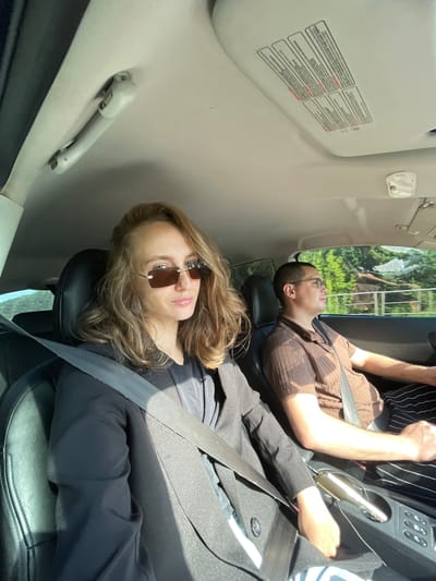 In Ludorvai, Russia, two individuals are seated inside a car. The female passenger, in the foreground and to the left, wears a black blazer over a black top and rectangular sunglasses. She has long, blonde-brown hair. The male driver, on the right, wears a brown, ribbed short-sleeved shirt and glasses. He is looking forward, with his hands on the steering wheel. The interior of the car is visible, including the ceiling with warning labels and a light fixture, the dashboard, and the center console. Outside the car, through the windows, trees and foliage are visible under a bright sky, suggesting a daytime scene. The overall impression is of a casual drive.