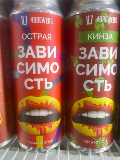 The image shows two cans of beer, likely sitting in a refrigerator or display case. The cans are red with a prominent graphic of a mouth with teeth, splattered with red liquid, resembling blood. Both cans feature large, white Cyrillic text that reads "ЗАВИСИМОСТЬ" (ZAVISIMOST), meaning "addiction." The top can has the word "КИНЗА" (KINZA), meaning "cilantro," on a green background, and the text "TOMATO GOSE." The bottom can has the word "ОСТРАЯ" (OSTRAYA), meaning "spicy," and also reads "TOMATO GOSE." The brand name "IV 4BREWERS" is visible on both cans. The scene appears to be indoors, with the cold, metallic structure of the display case visible. The lighting suggests it could be daytime, possibly in a store or bar. The overall aesthetic is bold and somewhat provocative, with the "addiction" theme and graphic imagery.