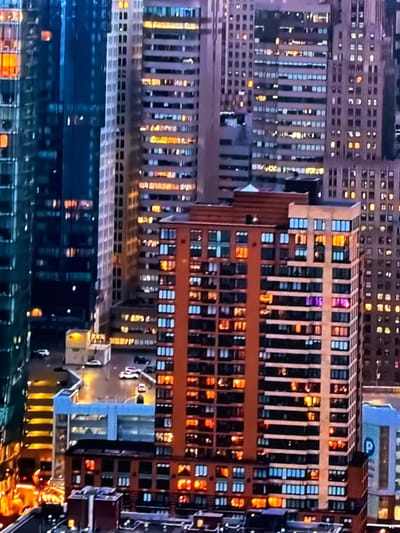 The image displays a dense urban cityscape at night or dusk, located in the city of Maidstone, United Kingdom. The scene is observed from an elevated viewpoint, likely from a tall building, looking across a concentration of high-rise structures. On the left, a dark, modern building with a blue-green glass facade reflects ambient light, with several internal windows illuminated in warm orange and yellow tones. To its right and in the background, numerous multi-story buildings of varying heights and architectural styles feature hundreds of windows, a significant portion of which are lit, casting warm yellow, white, and occasional blue lights. In the foreground-center, a prominent reddish-brown building, appearing to be residential or mixed-use, displays a grid of uniformly spaced windows. Many of these windows are illuminated with warm orange light, while some on the upper levels show distinct blue and magenta or purple light. Below the primary buildings, sections of ground level are visible, showing what appear to be streets or parking lots with several vehicles, primarily white and dark sedans, either parked or in motion. Small ground-level structures and illuminated streetlights are also present. A blue sign with a white 'P' symbol, indicative of a parking facility, is visible on the bottom-right. No discernible people, direct actions, or interactions are visible within the frame.