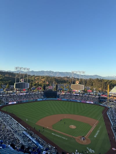 The image depicts a high-angle view of Dodger Stadium, a baseball park in Los Angeles, California, United States. The field is a verdant green with visible line markings and base paths. The stands are populated with spectators, primarily wearing blue, suggesting a home game. Two large scoreboards are positioned beyond the outfield fence, displaying the number 76. A backdrop of green trees and distant mountain ranges is visible behind the stadium. The sky is clear and blue.