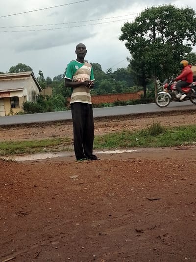 A lone man in a green and white striped t-shirt and black pants stands in a dirt lot in Kivaywa, Kenya. He stands facing the camera with his hands clasped in front of him. The man is standing to the right of a paved road.  A motorcycle is visible in the background, heading in the direction of the man. There are two buildings visible in the background, as well as several trees. The sky is overcast and a small puddle of water is visible in the dirt lot. The coordinates (latitude: 0.56, longitude: 34.806) place the location in the town of Kivaywa, Kenya.