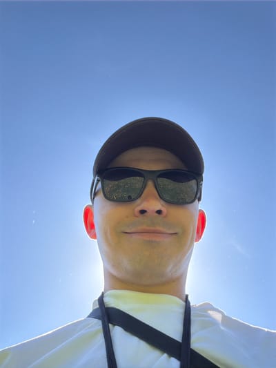 A low-angle close-up shot of a person wearing a baseball cap and sunglasses, set against a clear blue sky. The person appears to be wearing a white shirt with a black harness strap across the chest. The reflection in the sunglasses lenses is indistinct, but it suggests an outdoor environment. The skin tone is light, with noticeable redness in the ears. The photograph does not provide enough information to confirm the location is Oreshniki, Russia. No specific actions or interactions are visible.