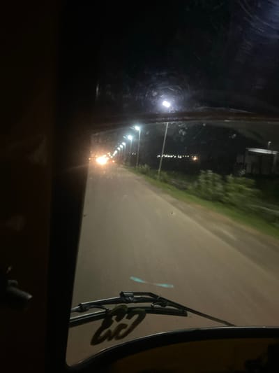 This media file captures a nighttime scene from the perspective of someone inside a vehicle, likely an auto-rickshaw or similar transport, looking out of a window. The primary focus is a road illuminated by a series of streetlights, creating streaks of light due to the low-light conditions and potentially a moving camera. The road itself appears to be paved and curves to the right, with a grassy or vegetated area lining the left side. In the distance, several more streetlights are visible, suggesting an urban or semi-urban environment.

On the right side of the frame, a portion of the vehicle's interior is visible, including a rearview mirror. The mirror reflects a stylized graphic or logo, which appears to be text written in a dark color with a green outline. The letters "CU" are clearly discernible, followed by what looks like "Y" or another cursive character. The time of day is clearly night, as indicated by the artificial illumination and the darkness of the surroundings. There are no visible people, but the scene suggests a journey is underway. The location is identified as Kuson, Nigeria. The overall mood is somewhat utilitarian, with the focus on the journey through the dimly lit landscape.