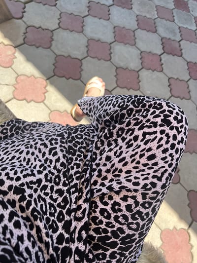 A close-up, eye-level shot captures a person's legs and part of their torso, clad in black and white leopard-print pants. The person is wearing light-colored sandals, and one foot is visible. The background consists of outdoor paving stones with a decorative pattern of light gray and pink. Sunlight casts shadows across the paving stones, creating a dappled effect. The image appears to be taken outdoors in an urban environment.