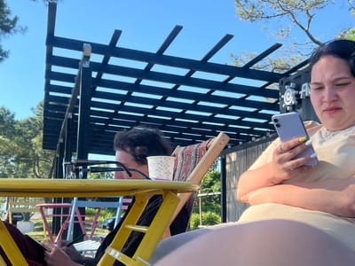The image shows two people sitting outdoors under a dark pergola. The person on the right is a woman with dark hair and a nose ring, holding a light purple smartphone. She is wearing a light-colored shirt and has brightly colored fingernails. The person on the left is partially obscured by a yellow table and appears to be writing in a notebook. They are wearing glasses. In the background, there are trees, a building with a logo, and colorful chairs and tables, suggesting a cafe or outdoor seating area. The overall scene is brightly lit by sunlight. The location is identified as the city of Ochamchire, Abkhazia.
