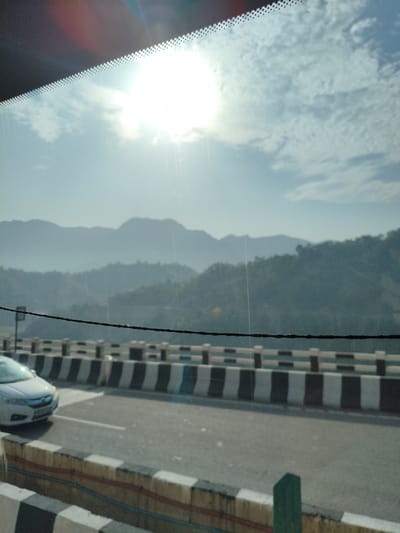 A bright, sunlit daytime scene shows a view through a windshield of a car driving on a highway. The sun is high in the sky, with some clouds visible. In the background, rolling hills and mountains are visible, shrouded in a light haze. A highway bridge with black and white striped guardrails is in the foreground, with a white car driving on the road. A black wire runs horizontally across the frame, likely part of the car's wiper system or an overhead line. The scene is likely somewhere along the National Highway 307 in or near Bilaspur, Himachal Pradesh, India, given the mountainous terrain and highway infrastructure.