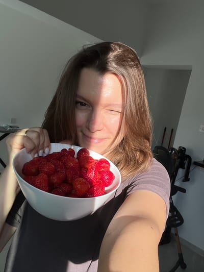 A woman is holding a bowl of strawberries. She is winking at the camera. The image is taken indoors, with sunlight streaming in from the right side. The woman is wearing a grey t-shirt. The background is out of focus but appears to be a room with some exercise equipment. The location is identified as Limassol, Cyprus.