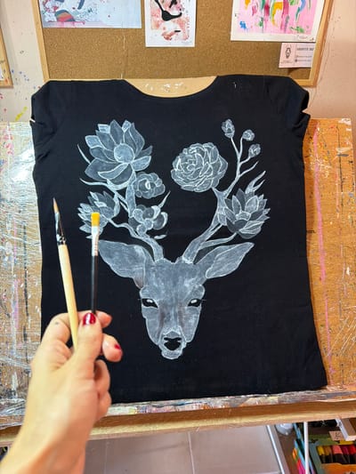 The image shows a black t-shirt with a white painted design of a deer's head adorned with flowers. The t-shirt is laid flat on a wooden surface, possibly an easel or workbench, which is splattered with various colors of paint. Two paintbrushes are held in a person's hand, which is visible in the lower left corner of the image. The hand has red nail polish. In the background, there is a corkboard with several framed artworks pinned to it. The overall setting suggests an art studio or creative space. There is no specific information in the image to indicate the location in Svilengrad, Bulgaria.