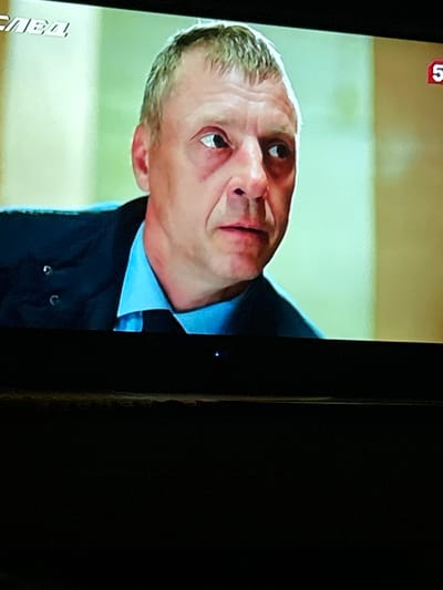 The image is a close-up shot of a middle-aged Caucasian man's face and upper body, taken from a television screen. The man has fair skin, thinning gray hair, and appears to be wearing a dark suit jacket over a light blue collared shirt. His expression is one of surprise or concern, with his eyes wide and his mouth slightly open. There are visible signs of fatigue or stress around his eyes.

In the upper left corner of the screen, there is text in a foreign language, possibly Russian, which is partially obscured. In the upper right corner, a red graphic with the number "5" is visible. The background is blurred and appears to be an indoor setting, possibly a hallway or room with a light-colored wall.

There is no identifiable information in the image that would allow for a specific location in Gelendzhik, Russia, or any other city to be determined. The image focuses solely on the man and the television screen he is being displayed on.