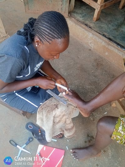 In Unguwan Rimi, Nigeria, a close-up, high-angle outdoor scene depicts a pedicure service in progress on a concrete surface. A dark-skinned female, identified as the pedicurist, is positioned on the left, crouched or seated, wearing a grey t-shirt with white lettering and dark lower garment, and has her hair styled in cornrows pulled back. She is actively filing the toenails of a client's right foot with a small white nail file. Her left hand holds a black smartphone. The client's lower right leg and foot are visible on the right, resting on a light-colored, patterned towel placed on the concrete. The client's toenails are painted white or have white tips applied. Scattered white nail tips, a larger grey foot file, a pair of black-handled scissors, a small red book or folder, and a small tube are visible on the concrete floor. A pair of dark, beaded sandals lies near the towel. In the background, a light brown wooden structure and a darker wall are partially visible. The scene is illuminated by natural light.