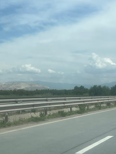This media file depicts a landscape viewed from what appears to be a moving vehicle on a highway during the daytime. The image, though presented vertically, captures a horizontal scene.

In the foreground, to the right, a grey road surface is visible, featuring a clear white lane marking. To the left of the road, a highway median or barrier system runs parallel, constructed from multiple horizontal metal rails or pipes, interspersed with small red reflective markers. The median area beneath the barrier shows sparse, dry-looking grass and small plants.

Beyond the median, a line of dark green trees forms a visual boundary, behind which the natural landscape unfolds. In the mid-ground, a large hill or mountain is prominent, characterized by exposed, light-grey rock faces, suggesting either natural erosion or possibly quarrying activity. Further in the distance, additional layers of hazy, muted blue mountains extend towards the horizon. The sky above is bright, with scattered white cumulus clouds against a lighter blue backdrop, indicating fair weather.

There are no people or explicit activities visible. No text is discernible in the image. The scene suggests travel through a mountainous region, consistent with the provided location context of Ayntap, Armenia.