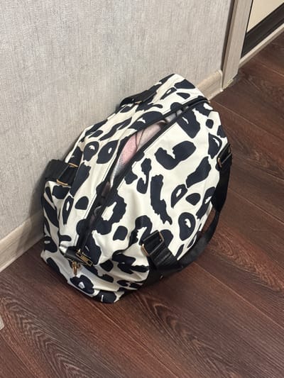 An open duffel bag with a black and white abstract pattern sits on a wooden floor next to a grey, textured surface. The bag is partially filled with a pink fabric, suggesting it is packed or being packed. The bag has black handles and straps with gold-colored hardware. The wooden floor has a warm, brown tone with visible grain patterns, and it appears to be a plank floor. The grey surface could be a rug, a mat, or a different type of flooring. The scene is brightly lit, indicating daytime, and there are no visible weather cues. The setting appears to be indoors, likely a home or a storage area, given the flooring and the presence of the duffel bag. There are no people visible in the image, and no text is present. The overall impression is a simple, still-life shot of an object in a domestic environment.
