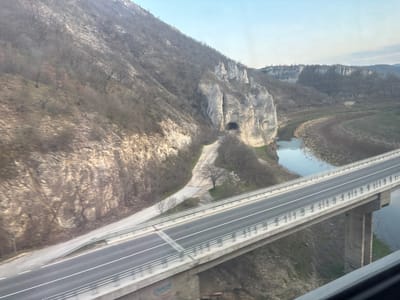 This is an elevated view of a river valley scene located in Struya, Bulgaria. A multi-lane concrete highway bridge, featuring grey guardrails and white lane markings, spans a winding river. Graffiti is visible on a concrete support column beneath the bridge deck on the left side.

To the left of the bridge, a steep mountain slope rises, covered predominantly with bare deciduous trees and rocky terrain. A dirt path runs along the base of this slope, leading towards the river. On the right bank of the river, a large, prominent limestone rock formation features a dark, oval-shaped cave entrance. The river itself is calm, reflecting a pale blue sky, and its banks display a mix of sparse green vegetation and brown earth. A solitary, bare deciduous tree stands near the dirt path close to the river. The distant background shows further hilly terrain and rock formations. No individuals or vehicles are present, and no specific actions or interactions are depicted. The environment suggests a transitional season, such as late autumn or early spring, due to the lack of foliage on most trees.
