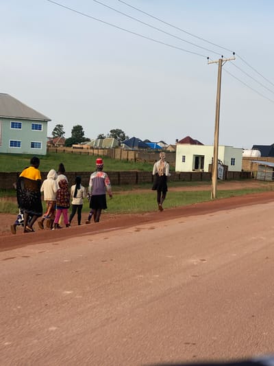 In the city of Kwang, Nigeria, a group of seven individuals, appearing to be women and children, walk along a dirt road. The group is moving from left to right in the frame. The person at the front of the group on the left wears a bright yellow top and a patterned black wrap. The individuals behind her are dressed in various colorful clothing, including a white hoodie, patterned pants, and a vibrant red and white head covering. The person furthest to the right in the group wears a patterned jacket over a darker skirt.

In the background, several buildings are visible. A light green, two-story building with blue-trimmed windows is on the far left. Other buildings, some with dark tiled roofs and some with lighter colored walls, are scattered throughout the scene. A wooden utility pole with power lines extends from the right side of the frame towards the center. The ground is predominantly a reddish-brown dirt, with patches of green grass appearing between the road and the buildings. The sky is a pale blue with a hazy appearance. The lighting suggests it is either early morning or late afternoon.