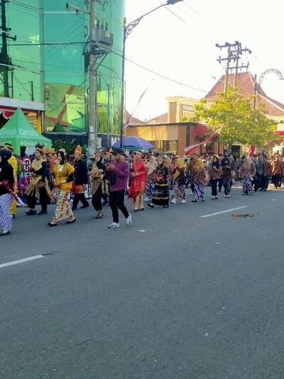 This image captures a daytime cultural procession occurring on an asphalt street in Madiun, Indonesia. Approximately 30-40 individuals are visible, walking in a line from left to right. The majority are adorned in varied traditional Indonesian attire, including batik sarongs, elaborate kebayas, and decorative headpieces. A male participant in the mid-foreground wears a purple long-sleeved shirt, black trousers, and white athletic shoes, holding a smartphone.

To the left, a tall building with green reflective glass dominates the background, with a green "Grab" branded tent on the sidewalk below it. An electrical pole with a transformer stands prominently in front of this building. To the right, various buildings are present, one featuring a distinct red-tiled traditional Indonesian roof. A tree with green foliage partially obscures the background, and red and white banners are strung between utility poles, indicative of national celebration. Utility wires and a decorative streetlamp are visible overhead against a bright sky.