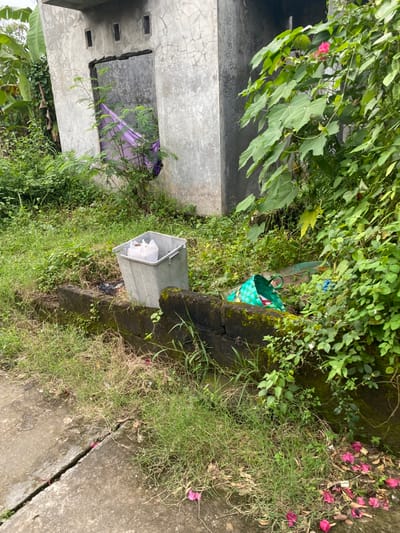 The image depicts a concrete wall, a metal trash can, and a green woven basket. The trash can is partially filled with a white plastic bag. The woven basket is near the wall and the trash can, with the green side facing the viewer. The scene is located in the city of Kecamatan Piyungan, Indonesia, based on coordinates (latitude: -7.827, longitude: 110.481). The scene is outdoors, with green foliage and a concrete path in the foreground. The wall is covered with moss. There are several pink flowers scattered on the ground. There are no people present in the image.