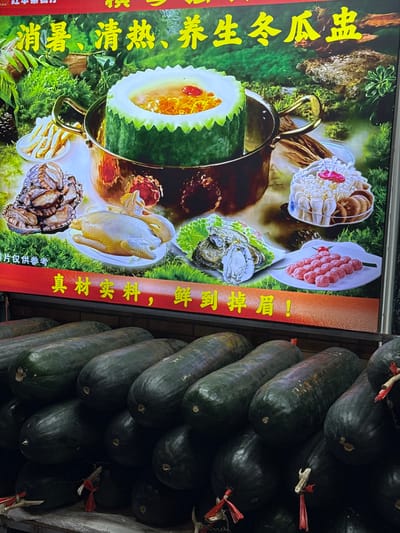 A close-up shot reveals a sign advertising "Winter Melon Soup" with images of various ingredients, including fresh seafood, chicken, and mushrooms. Beside the sign are a large number of winter melons stacked on top of each other. The sign features Chinese characters that translate to "Summer heat relief, clearing heat, nourishing winter melon soup," and a note that "pictures are for reference only." It also says: "real material, fresh to eyebrows falling." The sign is advertising this soup in Xiangzhou, China.