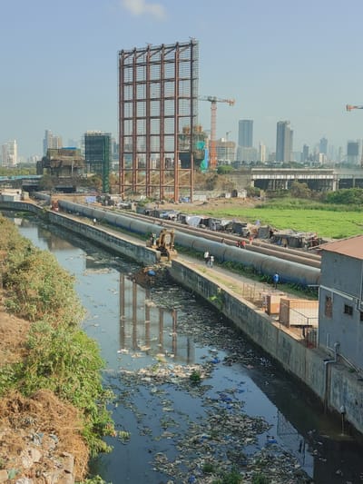 The image depicts a polluted canal in Mumbai, India. The canal is filled with trash and debris, and its water appears dark and stagnant. Along one side of the canal, large industrial pipes run parallel to the waterway. Makeshift structures, possibly temporary housing, are built on top of these pipes. Several people are visible walking on or near the pipes, some near an excavator. In the background, a large metal framework structure and other buildings are visible, indicating an urban environment. Construction cranes are also present in the background. The sky is clear and bright. Overall, the scene shows a stark contrast between urban development and environmental degradation.
