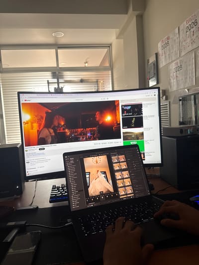 The image shows a workspace with two monitors displaying content on YouTube and a photo editing program. A person's hands are visible on a keyboard, suggesting interaction with the computer. Calendars for the year 2026 are attached to the wall. There is also an audio interface from MAudio, and another keyboard. The window shows a bright, sunlit space but no details that point specifically to Cholula, Mexico. There is a phone and ledger sitting on the table.