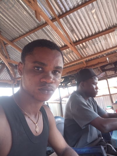 The image captures two males within a semi-enclosed structure in Omicha Umu Awada, Nigeria. In the immediate foreground, a young adult male with dark skin and short dark hair looks directly at the camera with a neutral expression. He wears a black sleeveless top and a gold-colored chain necklace with a quatrefoil pendant. To his right and in the mid-ground, a second young adult male with dark skin is partially visible, facing towards the right side of the frame. This individual wears a grey short-sleeved t-shirt and a dark baseball cap worn backward, and appears to be seated on a blue plastic chair, with his right arm resting on a light blue surface. The overhead structure consists of a corrugated metal roof supported by wooden beams and rafters, suggesting a basic, open-air or partially covered setup. Natural light illuminates the scene.