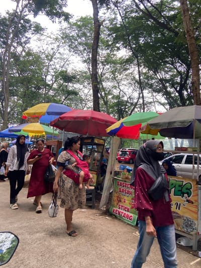 A street scene with several people and food stalls under colorful umbrellas. A woman carries a baby in a carrier. Another woman, wearing jeans and a maroon top, walks towards a food stall selling snacks like "Basreng" and "Stick Kentang". Several other individuals stand nearby, some wearing hijabs. A car is visible in the background. Trees are visible on the side of the road.