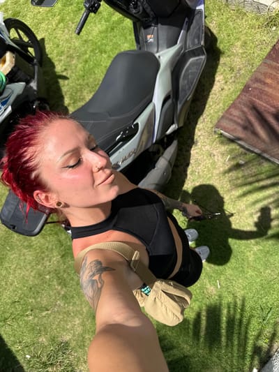 A medium shot of a red-haired person wearing a black top, black shorts, and gray sneakers standing on a green lawn. The person is adjacent to a parked motorcycle. The location is identified as Dalung, Indonesia.
