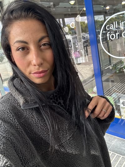 A selfie of a woman with dark hair, brown eyes, and light-pink lipstick, wearing a grey, fleece-textured jacket with a black, animal-print lining visible near the zipper. She has a silver ring on one of her fingers. The background appears to be a storefront window with a sign that reads "call in for". Reflected in the glass are objects and vehicles indicating an urban environment, likely the city of Warrington, United Kingdom. The interior details behind the glass are not discernible.