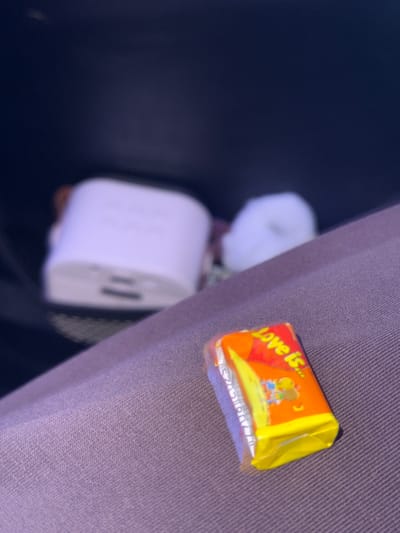 The image, taken with a slight tilt, displays several objects in what appears to be an indoor or vehicle setting in Klenovskoe, Russia. The most prominent item is a "Love is..." candy or gum wrapper, positioned in the upper right quadrant on a textured, purplish-grey fabric surface. The wrapper is primarily orange and yellow, featuring a graphic of a boy and a girl. The text "Love is..." is clearly visible on the wrapper.
To the left and slightly out of focus, a white, cylindrical electronic device is present, possibly a power bank or charger, with what appear to be USB ports on one end. Another blurry white object is partially visible behind it. The background on the left is dark and indistinct, contrasting with the lighter fabric surface that occupies the right side of the frame. No people or specific activities are depicted. The lighting is dim, suggesting an indoor environment or nighttime conditions.