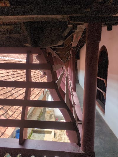 This image depicts a covered wooden walkway within the Mattancherry Palace in Mattancherry, Kochi, India, viewed from an elevated perspective. The foreground features a reddish-brown wooden balustrade with carved detailing along its lower edge. To the left, a portion of a sloped roof covered with terracotta tiles is visible. Below the walkway and roofline, an open-air concrete courtyard or alleyway is observable, containing some vegetation and a blue cylindrical barrel. The right side of the image is dominated by a long, colonnaded walkway, supported by multiple reddish-brown wooden pillars. This walkway runs parallel to a white-painted wall that features at least one dark, arched opening. The ceiling structure above the walkway consists of dark, exposed wooden beams and supporting elements. The scene is illuminated by natural light. No individuals are depicted.