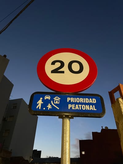 This image captures a street sign in La Línea de la Concepción, Spain, at dusk. The primary subject is a circular speed limit sign displaying "20" within a red border and white background. Below it, a rectangular blue sign reads "PRIORIDAD PEATONAL" (Pedestrian Priority) and features white pictograms of a car, a house with a ramp, and two people, one adult and one child, walking. The signs are mounted on a metal pole. In the background, tall, modern buildings with visible windows are silhouetted against a darkening blue sky. A single power line runs across the top left of the frame. The scene appears to be an urban street setting.