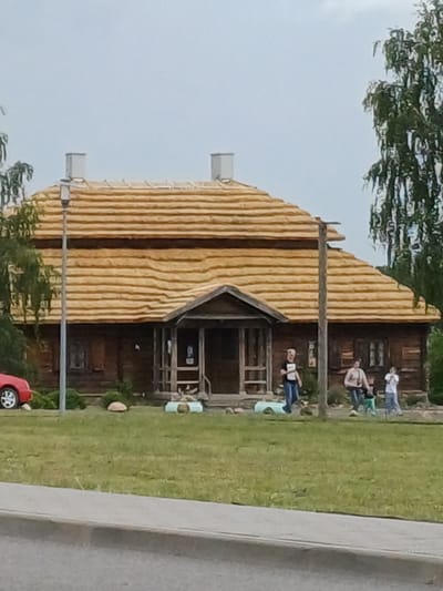 The image depicts a wooden house with a thatched roof and two chimneys, located in Квасевіцкі сельскі Савет, Belarus. In front of the house, a red car is parked on the left side. Several individuals are present on the lawn, including one person walking a dog and another person standing near the entrance. The environment is a grassy area with a paved walkway in the foreground. The sky is overcast.
