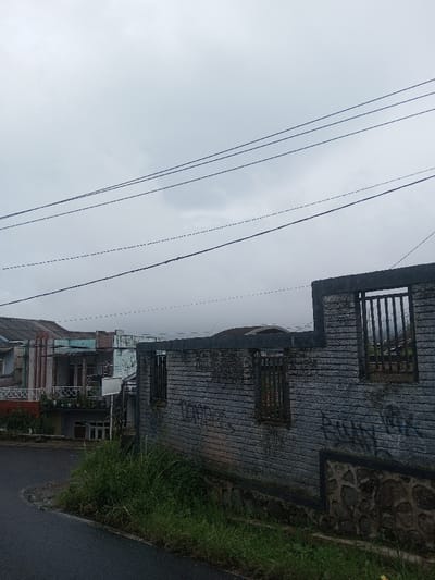A street scene is captured under an overcast sky. A worn, grey brick wall with three window openings stands beside the road. Patches of green grass grow at its base. In the background, residential houses with balconies and unique architectural details are partially visible. Power lines stretch across the cloudy sky, adding to the overall atmosphere of a somewhat bleak day.