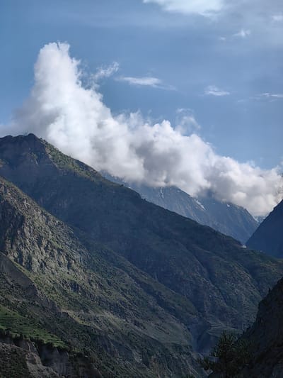 The image captures a breathtaking view of the Himalayan mountains near Kardang, India. The foreground is dominated by steep, rocky slopes covered with patches of green vegetation and scattered trees. The mountains extend into the background, with some peaks showing remnants of snow. A dramatic cloud formation drifts across the sky, partially obscuring some of the higher mountain ranges. The sky is a clear blue, with a few wispy clouds in addition to the large cloud mass. The overall impression is one of rugged, natural beauty and solitude. There are no people or animals visible in the image. A small, white structure is visible on one of the distant slopes, likely a dwelling or a religious site. The lighting suggests it is daytime, possibly late afternoon, given the shadows cast on the mountains.
