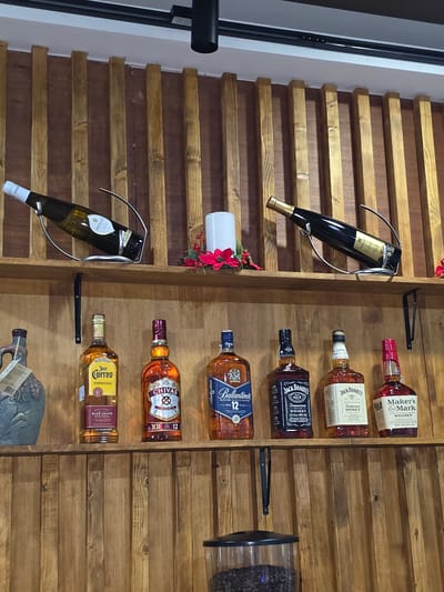 The image depicts a wooden shelf displaying various alcoholic beverages. On the top shelf, there are two wine bottles, one white and one dark, each supported by metal stands. To the right of the wine bottles, a white candle sits next to a red floral arrangement. The lower shelf holds a collection of liquor bottles. From left to right, these include a Jose Cuervo Especial tequila bottle, a Chivas Regal 12 Year Old Scotch whisky bottle, a Ballantine's 12 Year Old Blended Scotch Whisky bottle, two Jack Daniel's Tennessee Whiskey bottles (one Old No. 7 and one Tennessee Honey), and a Maker's Mark Kentucky Straight Bourbon Whisky bottle. To the far left of the lower shelf is a decorative stoneware jug. The background consists of vertical wooden slats. Below the lower shelf, a portion of a coffee grinder filled with coffee beans is visible. The overall setting appears to be a bar or a display area within a commercial establishment. The location is Asan-si, South Korea.