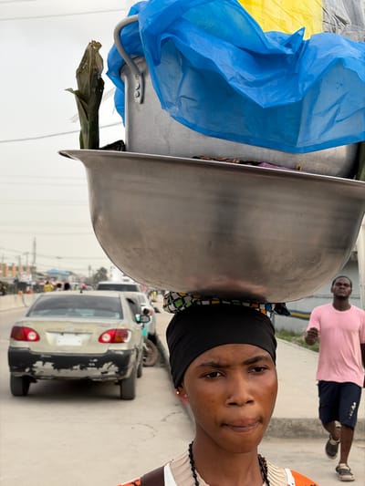 In Jakande, Nigeria, a young individual, likely female, is captured in a close-up, foreground view, carrying a substantial load on her head. She wears a black headwrap over a patterned fabric pad, supporting a large, reflective metallic basin. Stacked within or on top of the basin is a smaller aluminum pot, partially draped with bright blue and yellow plastic sheeting. A dark green, possibly leafy, item protrudes vertically from the contents. She has a subtle nose piercing, a small gold earring, and a dark beaded necklace. Her gaze is directed slightly to the left of the frame, with a neutral expression. In the mid-ground on the right, a male figure in a pink t-shirt and dark shorts walks. On the left, a light-colored sedan with visible paint damage on its rear bumper is parked or moving slowly on a paved road, which extends into the blurred distance with other vehicles and figures under an overcast sky. The scene depicts an urban street environment.