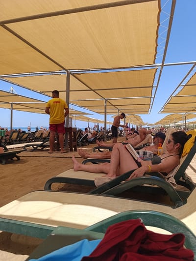 This is a medium-angle shot of a beach resort on a sunny day. The foreground features a number of empty, reclined beach chairs, some covered by a beige tarp overhead, and a few with towels and clothing draped over them. In the mid-ground, several people are relaxing on lounge chairs under large, beige tarp shades supported by metal poles.
The activity is leisurely relaxation at a beach. One woman in a bikini is reading a book on a lounge chair, while a man next to her, wearing a blue cap, is also reclining. Further back, a lifeguard in a yellow shirt with the word "LIFEGUARD" printed on the back and red shorts stands near a wooden structure. Other beachgoers are visible in the background, some wading in the ocean or playing volleyball.
The scene is set on a sandy beach with the ocean visible in the distance. The sky is clear and blue, indicating good weather. The lighting suggests it is daytime, likely late morning or early afternoon, given the angle of the sun and the bright illumination. The overall mood is one of relaxation and vacation. The location is identified as Manavgat, Turkey.