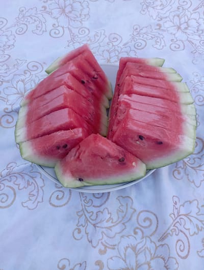 The image shows a close-up, overhead view of sliced watermelon on a white plate. The watermelon is cut into wedges, with the pink flesh and green rind visible. Some slices are stacked neatly, while others are slightly askew. The plate is placed on a white tablecloth with a subtle floral pattern in a gold or light brown color. The lighting suggests it is daytime, likely outdoors or in a well-lit room, with soft shadows. The setting appears to be a domestic or casual outdoor space, possibly a picnic or a meal. There are no people visible in the image. The scene evokes a sense of freshness and summer refreshment. The location context provided is Beruniy, Uzbekistan, which might suggest a regional style of presentation or local ingredients, though this is not explicitly evident from the image itself.