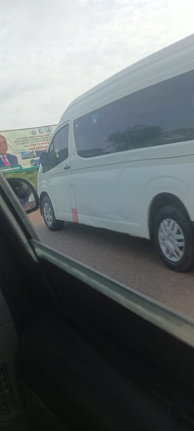The image shows a white van on a paved road in Opi, Nigeria. A political banner is visible in the background. The photo was taken from inside another vehicle.