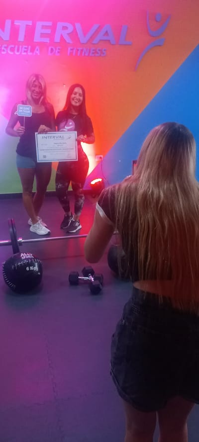 The scene depicts an indoor fitness facility, identified as "INTERVAL ESCUELA DE FITNESS," located in Quilmes, Argentina. Three women are present within a brightly lit area featuring a color-blocked wall (orange and blue) and a purple interlocking foam mat floor.
In the mid-ground, two women are posing. The woman on the left, with blonde hair, wears a black tank top, light-colored shorts, and white sneakers. She holds a white speech bubble sign stating "ME GANÉ UN TALLER" (I won a workshop). The woman on the right, with dark hair, wears a dark t-shirt with a logo and patterned dark leggings with sneakers. She holds a white certificate or diploma, clearly showing "INTERVAL" at the top and text indicating "INSTRUCTORADO DE MUSCULACIÓN Y PERSONAL TRAINER." Both women are smiling and looking forward.
In the foreground, a third woman stands with her back partially to the camera. She has long blonde hair and is wearing a dark, possibly short-sleeved top with white trim and dark denim shorts.
On the purple floor, fitness equipment is visible: a black barbell with one plate, a black "ALLY SPORTS 6 Kg" medicine ball, and a pair of black hex dumbbells. The environment is illuminated by vibrant pink and magenta lighting, with a red light source positioned on the floor near the feet of the two background women.