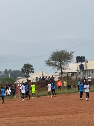 The image shows a dirt soccer field in Du, Nigeria, with a soccer match in progress. Several players are visible on the field, wearing different colored jerseys. Spectators are gathered behind a fence on one side of the field, and some are standing around the perimeter. In the background, there are buildings, trees, and a water tower. The sky is overcast, suggesting a cloudy day. The scene depicts a typical community soccer event.