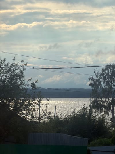 The image captures a tranquil waterscape possibly of a lake or wide river on an overcast day. Light glints off the water's surface. In the foreground, power lines are visible, obscuring the view of the body of water. Silhouetted trees and vegetation surround the water. A dark, possibly metallic, structure is also situated on the right side of the scene. Given the prompt context, it is inferred that the location is near Kryukovo, Russia. The picture does not contain any people. The environment includes the water, shoreline, vegetation, sky, and electrical lines. There are no visible actions or interactions.