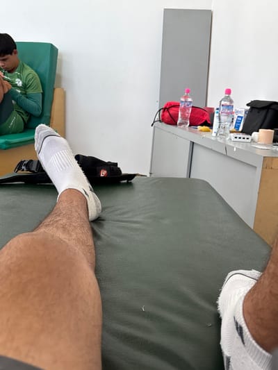 The image captures an indoor scene, likely within an athletic or medical facility. From a first-person perspective, the viewer's bare legs, clad in white athletic socks, rest upon a dark green padded surface, possibly an examination or treatment table. A pair of black sandals with a red logo is visible near the left foot. In the background, to the left, a young male individual in a green athletic long-sleeved shirt and shorts sits on a green cushioned bench, focused on a handheld electronic device. To the right, against a white wall, a light grey cabinet unit displays two clear plastic water bottles with pink caps, a red drawstring bag, and various items including white tubes, adhesive tape, a white multi-port electronic device, and a black pouch. The walls are white, and the lighting is even. There are no specific visual identifiers within the scene to confirm the location as Las Crucitas, Mexico.