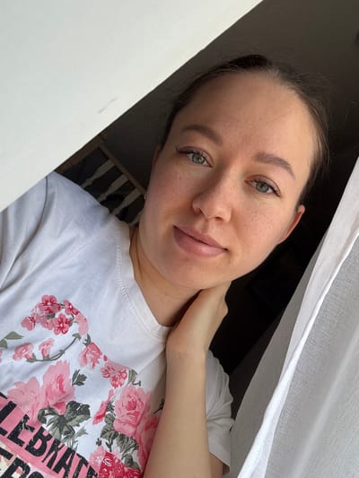 A light-skinned woman with freckles poses for a selfie. Her dark hair is pulled back, and she has green eyes and light pink lipstick. She is wearing a white t-shirt with a floral print. Her right hand is touching her neck. She is positioned in front of a white, translucent curtain and a window frame. The location is identified as Ферма, Russia.