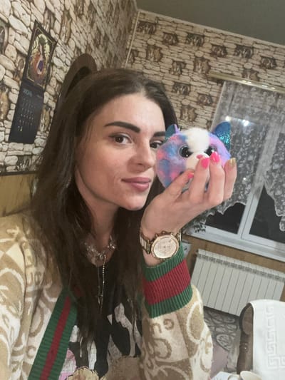 A young woman with long, dark hair is lying down, taking a selfie. She is holding a small, fluffy, multi-colored toy animal with blue glittery ears and big blue eyes up to the camera. Her left hand, with bright pink nail polish and one gold-glitter painted nail, is touching the toy. She is wearing a tan sweater with a pattern of interlocking beige letters and red and green stripes on the cuffs. A gold-colored watch with a white face and diamond embellishments is on her right wrist. A delicate silver necklace with a star pendant is visible around her neck.

The background appears to be an indoor setting. The wall to the left has a textured wallpaper resembling light brown bricks. A framed picture or mirror hangs on the wall. To the right, a white radiator is visible, and further back, a white patterned towel is draped over something brown.

The lighting suggests it is daytime, but no external windows are clearly visible to determine the exact time or weather. The woman's expression is neutral, with her eyes open and looking towards the camera.

On a surface below her, a small black calendar or booklet is laid open. The visible text on it includes the Russian word "июнь" (June). The overall scene is casual and personal. The woman is likely at home, given the indoor setting and personal items.