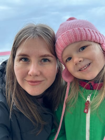 A close-up outdoor portrait taken in Gornyi (Dvurechenskoe S/P), Russia, features an adult female and a young child positioned side-by-side. The adult female occupies the left half of the frame, displaying light brown hair, light-colored eyes, dark eyebrows, and a small dark mole on her right cheek. She wears a dark-colored padded winter jacket and exhibits a slight closed-lip smile while looking directly at the camera. The young child, appearing to be a girl between 2-5 years old, occupies the right half of the frame. She wears a pink knitted winter hat with ear flaps and a top pompom, and a bright green winter jacket with a white zipper featuring a red 'R' logo pull tab. The child has blonde hair visible beneath the hat, light blue eyes, and a wide open-mouth smile revealing her upper and lower teeth, also looking directly at the camera. The background consists of an overcast, pale blue-grey sky. A building with a red roof is partially visible in the far left distance, and the ground beneath it appears light-colored.