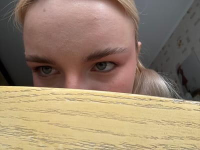The media file shows a close-up, upside-down view of a person's face peeking over a yellow surface. The subject is a person with blonde hair, and only their eyes, eyebrows, and forehead are visible. The yellow surface appears to be made of wood with a grain pattern. The background is out of focus and appears to be a room, possibly with wallpaper. The lighting suggests it is daytime. The person's expression is neutral, and they seem to be looking upwards. There is no visible text in the image. The provided location context is Sinyavino, Russia, which does not offer any specific visual cues within the image itself.