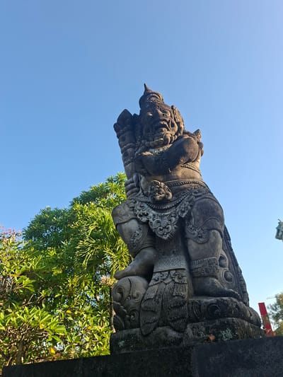 A low-angle, full shot captures a large, stone statue of a demonic figure against a clear blue sky. The statue is carved with intricate details, featuring a fierce expression, elaborate headdress, and ornate clothing. To the left of the statue, lush green trees with vibrant foliage fill the background, with hints of pink flowers visible. The statue is positioned on a dark, rough-textured pedestal, and a small patch of moss and a fallen leaf are visible on its surface. In the far right background, a distant red structure and a construction crane suggest an urban or developed area, potentially the city of Gianyar, Indonesia. The sunlight casts subtle shadows on the statue, highlighting its weathered texture.
